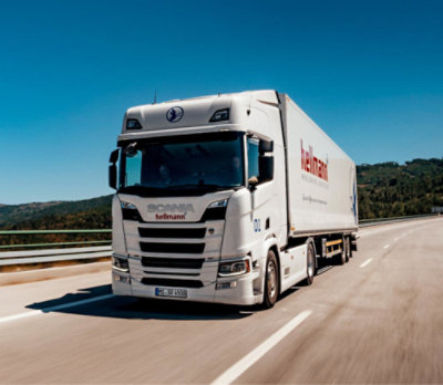 A white Scania semi-truck with "Hellmann" branding drives on a highway under a clear blue sky