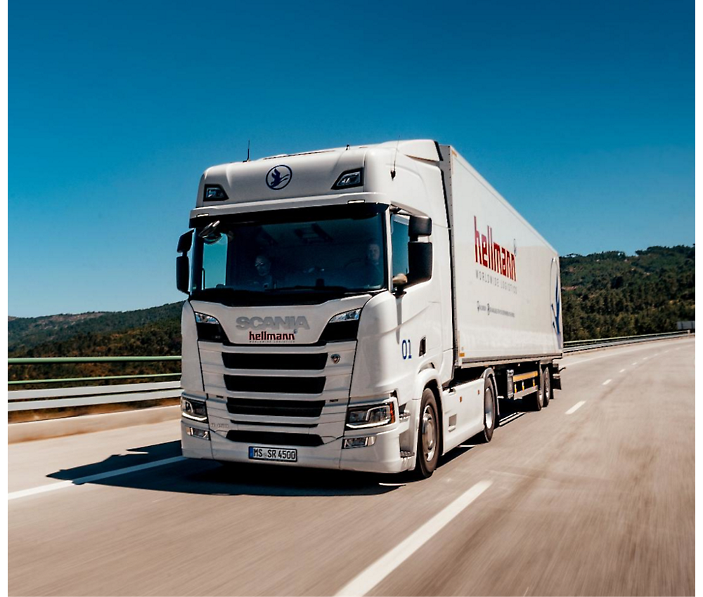 Um camião Scania branco com a marca "Hellmann" circula numa autoestrada sob um céu azul claro
