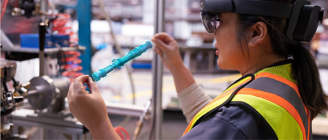 Een werknemer in een veiligheidsvest met een AR-bril onderzoekt een blauw mechanisch onderdeel in een industriële omgeving.