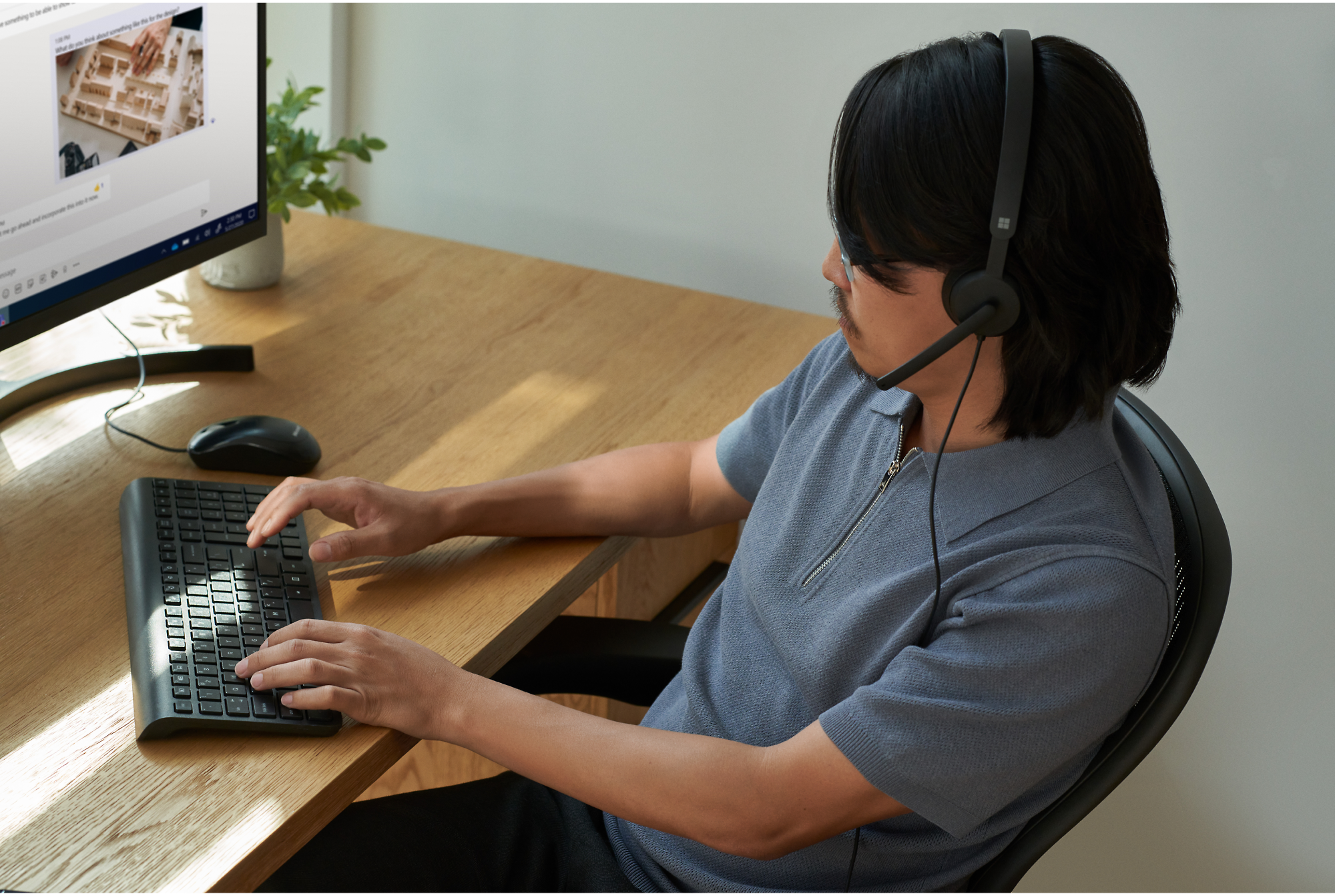 Kuulokemikrofonia käyttävä henkilö istuu pöydän ääressä, kirjoittaa näppäimistöllä ja katsoo tietokoneen näyttöä.