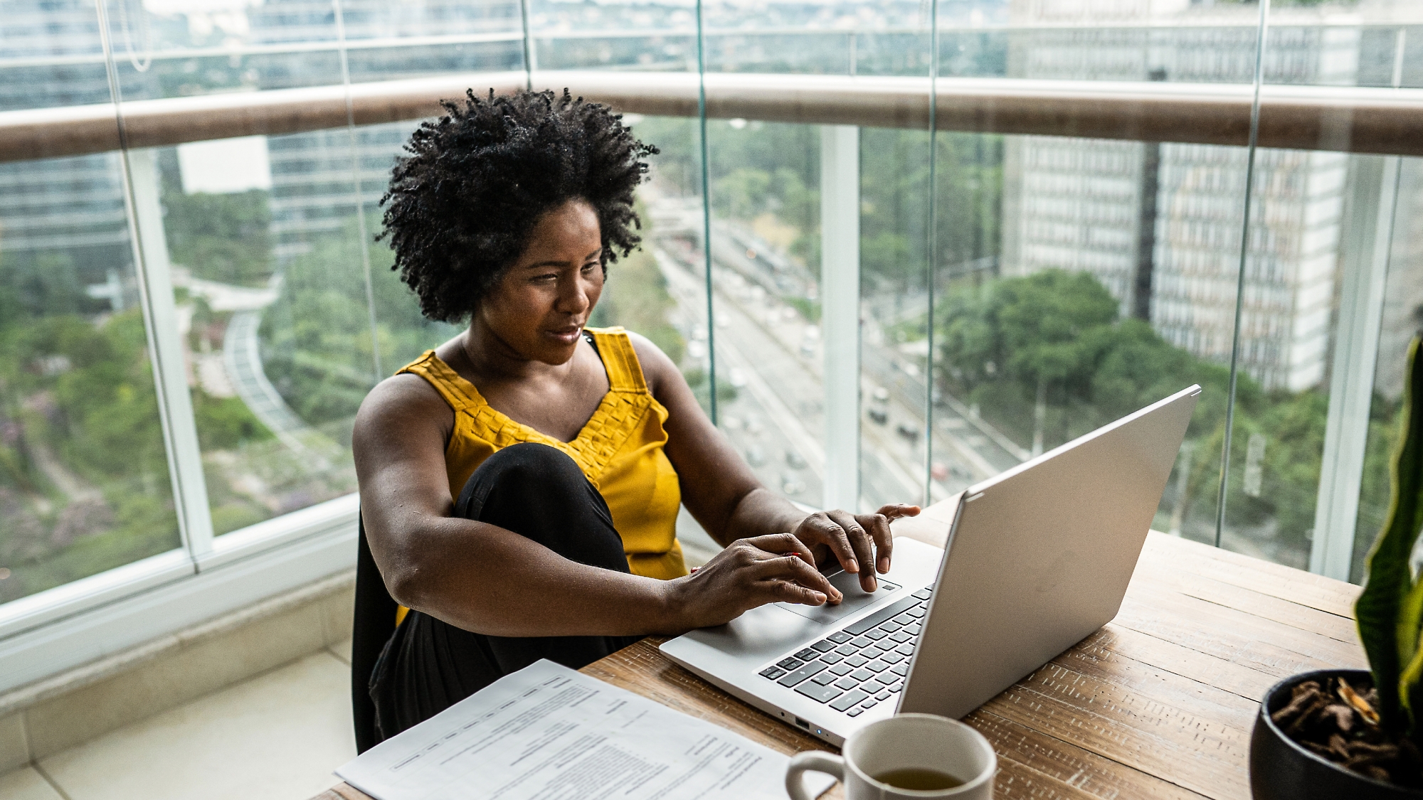 Une femme travaille sur un ordinateur portable à un bureau près d’une fenêtre qui donne sur un paysage urbain, avec des documents et une tasse de café à proximité.