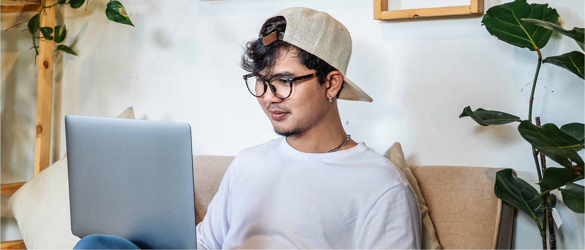 A person wearing glasses and a backward cap is sitting on a couch, using a laptop