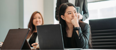 Two women sit at a conference table with laptops, listening attentively.