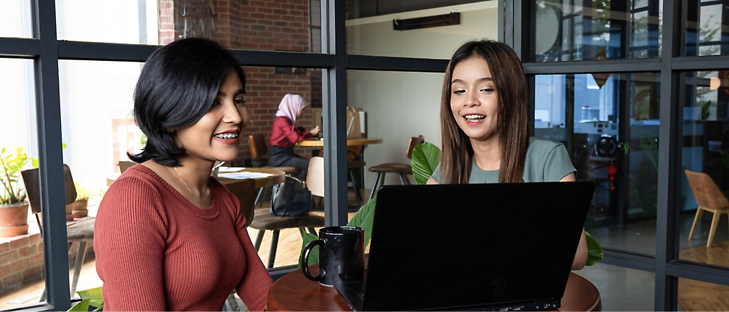 Dos mujeres sentadas en una mesa de despacho, mirando un ordenador portátil.Otras personas están trabajando en segundo plano.