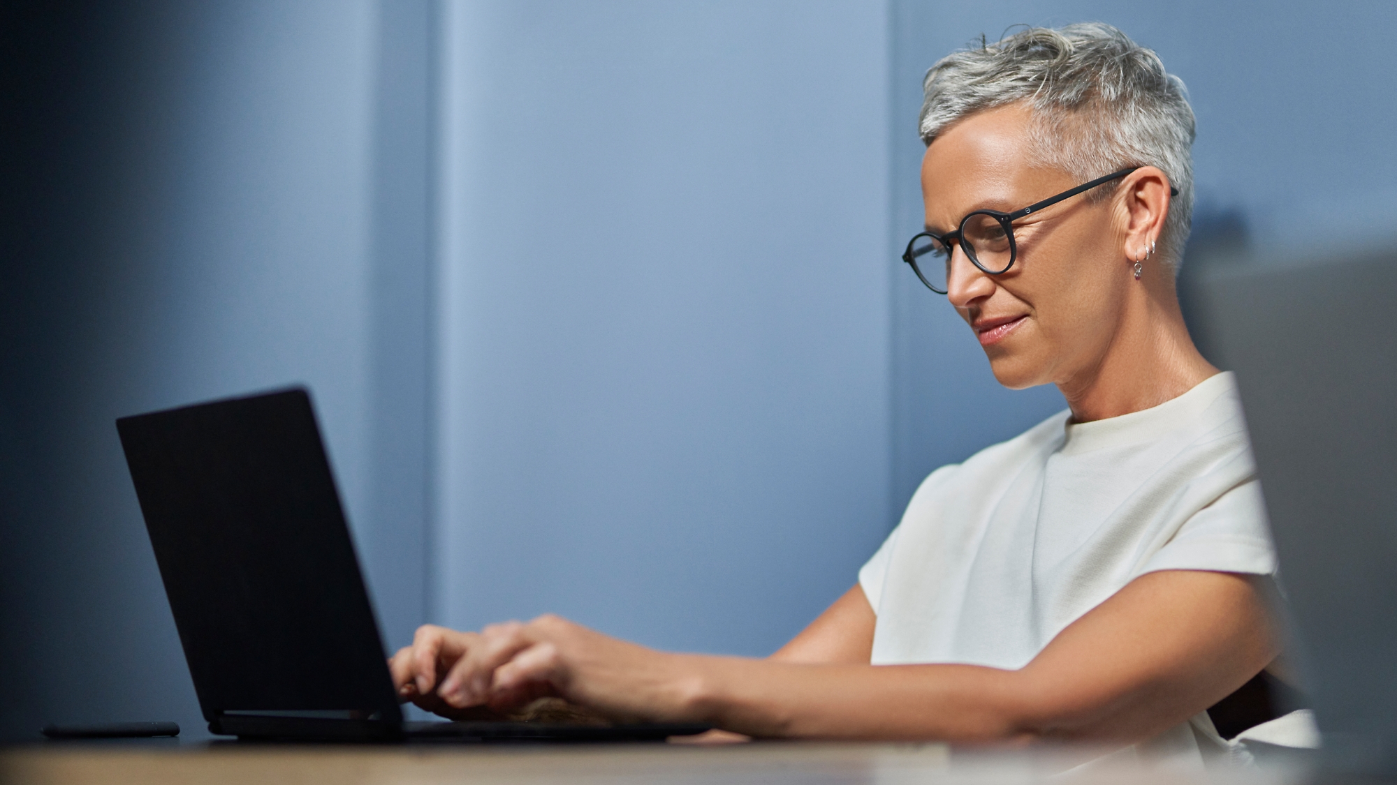 Dojrzała kobieta z krótkimi siwymi włosami uśmiechnięta i pracująca na laptopie w nowoczesnym biurze.