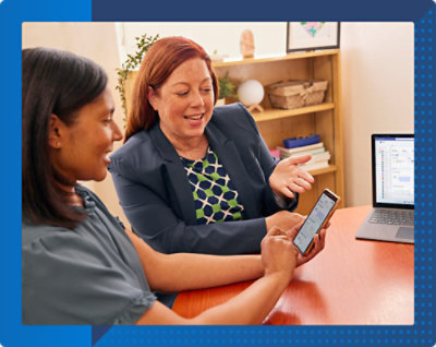 Two women sit at a table. One shows a smartphone screen to the other, who gestures while talking.