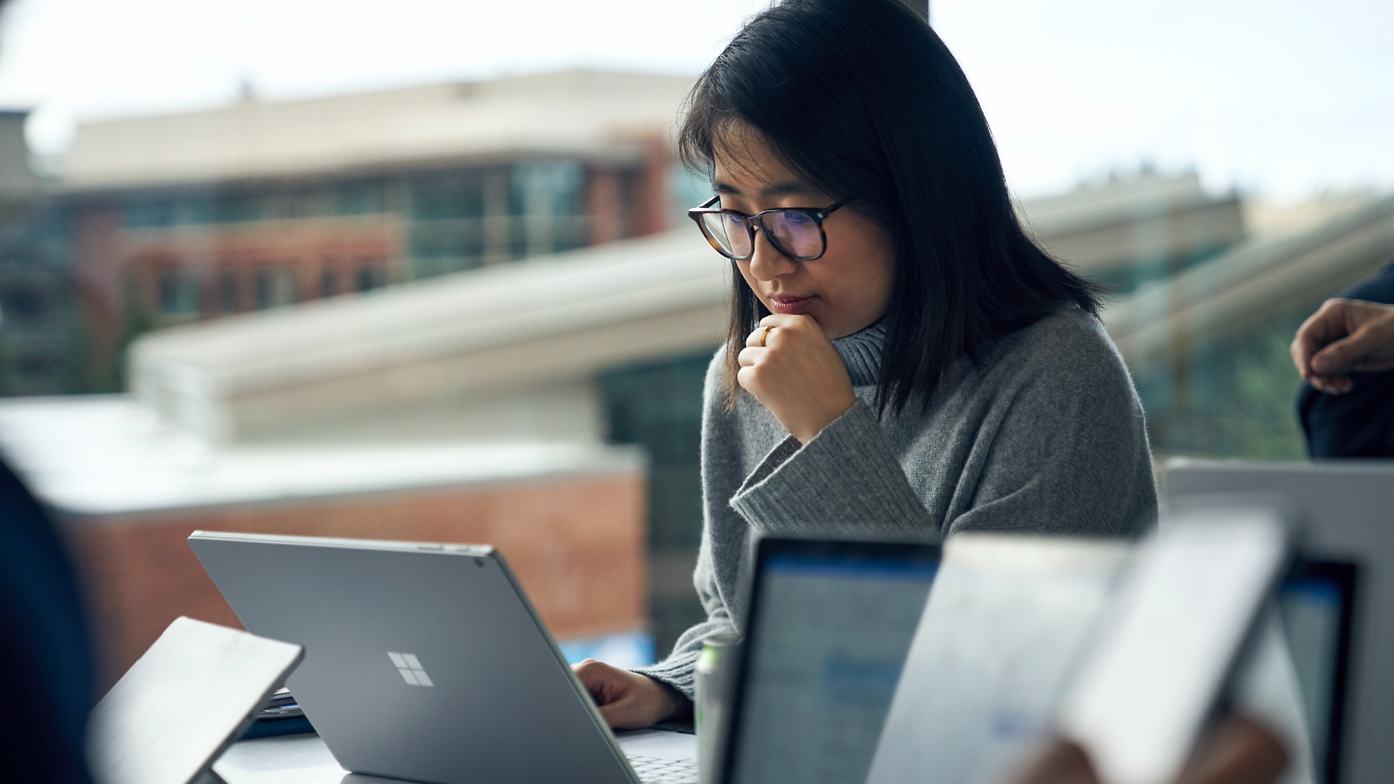 眼鏡をかけている集中しているアジアの若い女性は、他の従業員と一緒に、忙しいオフィスのテーブルでノート PC を使用します