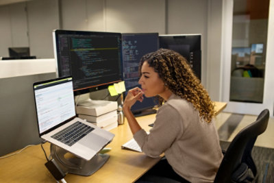 Woman sitting at a desk with multiple screens and looking at a laptop