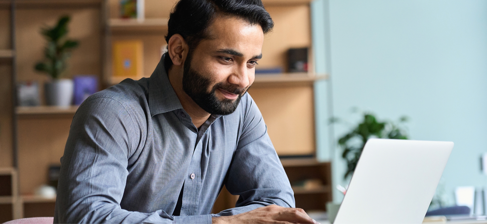 Een glimlachende man die op een laptop werkt in een kantoor met schappen op de achtergrond.