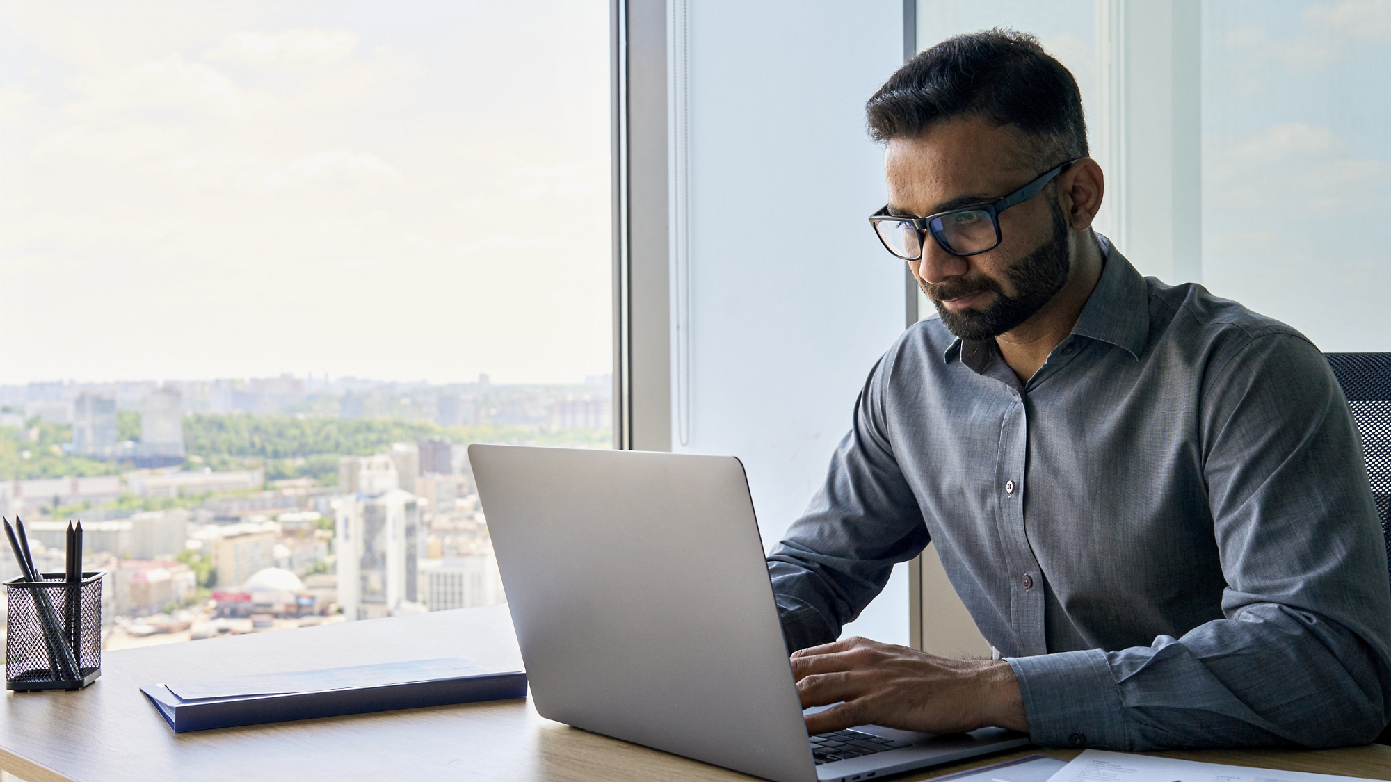 A man wearing glasses works intently on a laptop at a desk by a window overlooking a cityscape.