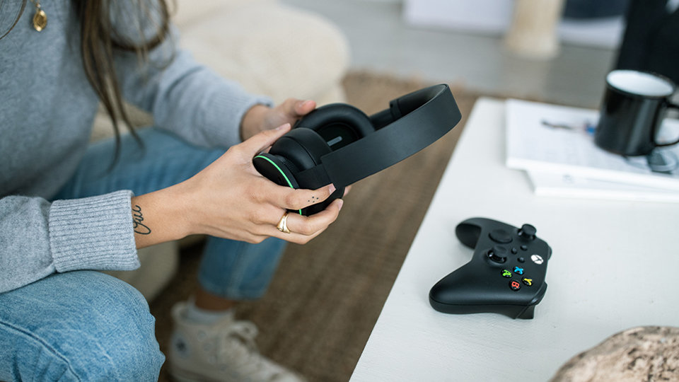 A person holds the Xbox Wireless Headset while sitting on a couch beside a coffee table.