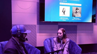 Two people with headsets in a recorded discussion with screen backdrop.