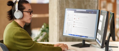 Une personne portant des écouteurs est assise à un bureau et utilise deux écrans d’ordinateur, dont l’un affiche une application de conversation instantanée.