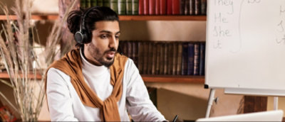 Person wearing headphones sitting in front of a laptop with a whiteboard and bookshelf in the background.