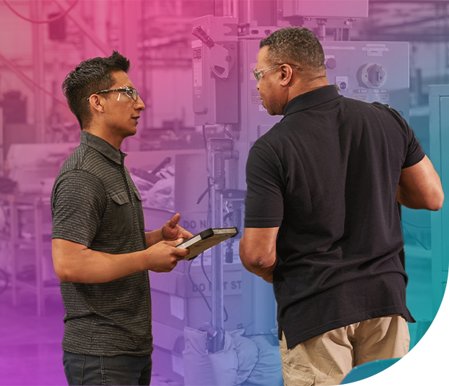 Two men in safety goggles and dark polos discuss equipment in front of machinery with a colorful light overlay. One holds a tablet-like device.