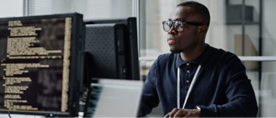 A man in glasses and a dark shirt works at a computer with lines of code on the screen in an office setting.
