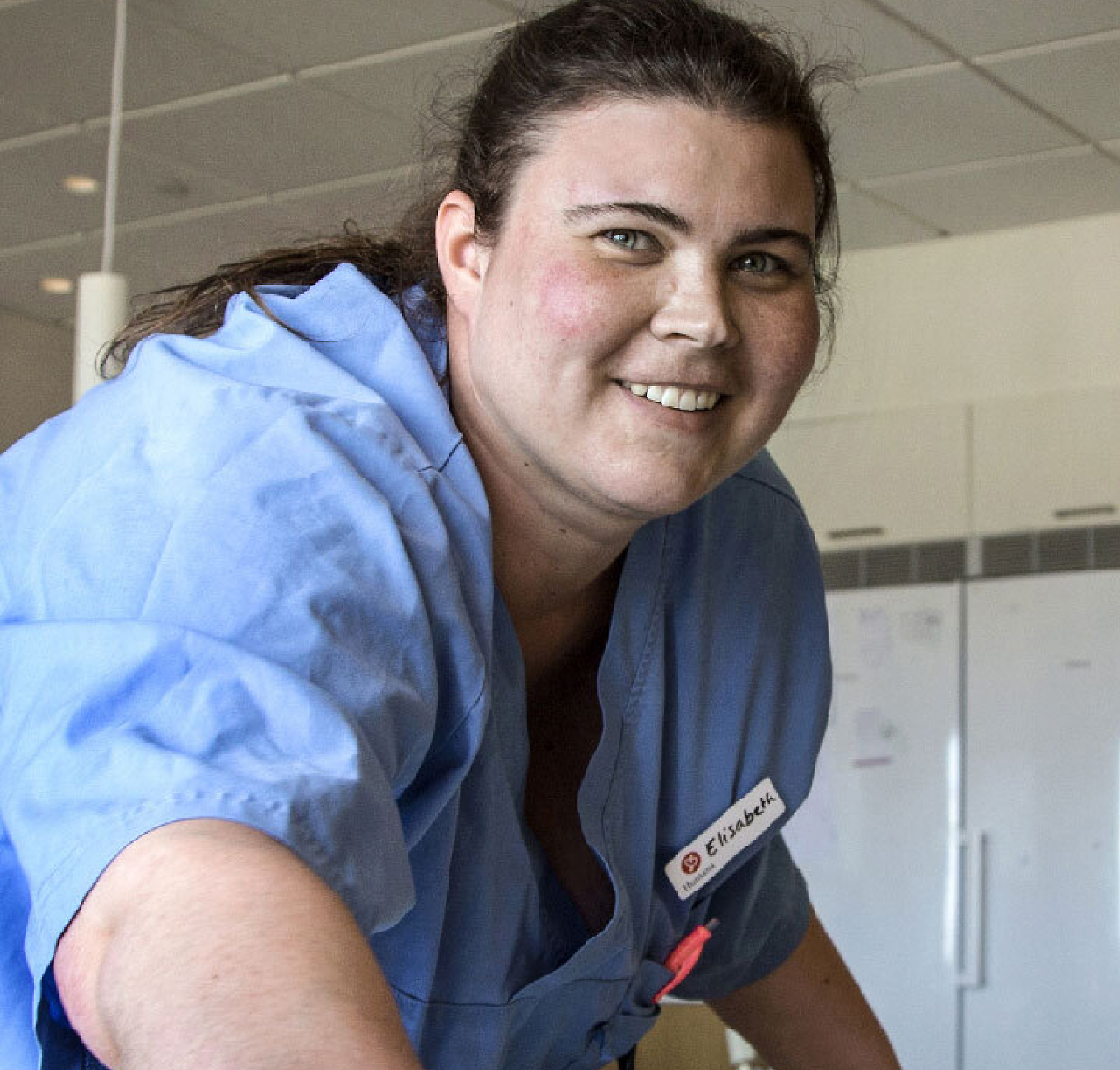 Một nữ y tá với một chiếc thẻ tên đánh dấu 'elizabeth' đang mỉm cười trong bộ đồng phục màu lam, trong không gian bệnh viện sáng và rộng.