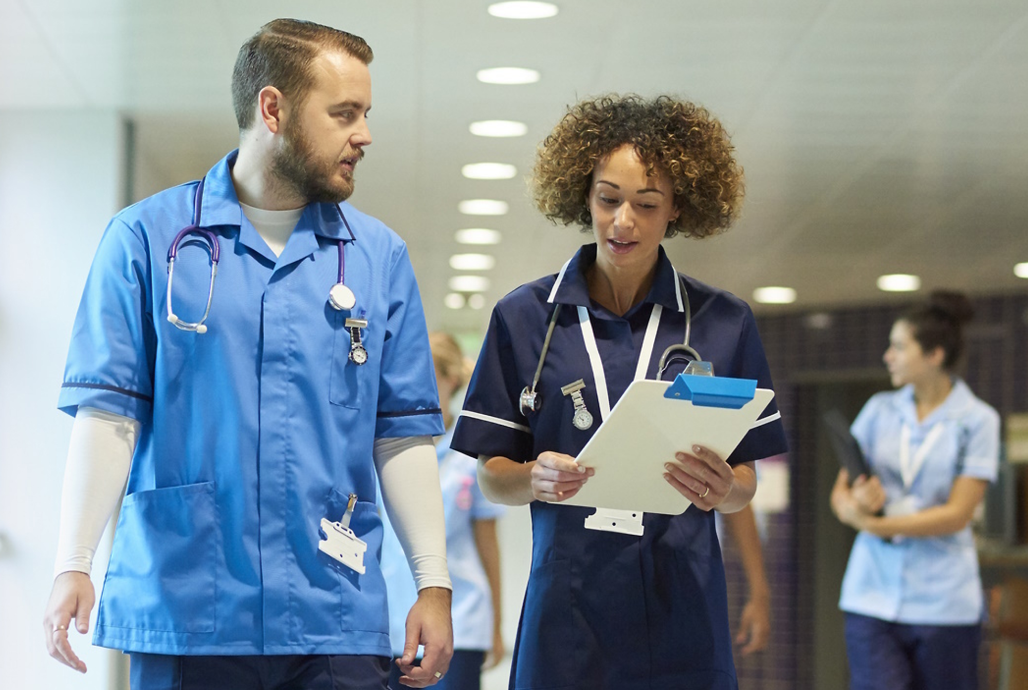 Dos profesionales sanitarios con uniformes azules hablando y caminando en un hospital, con uno sosteniendo un portapapeles. 