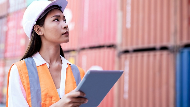 A female engineer wearing a helmet and reflective vest holds a tablet, standing in front of colorful shipping containers.