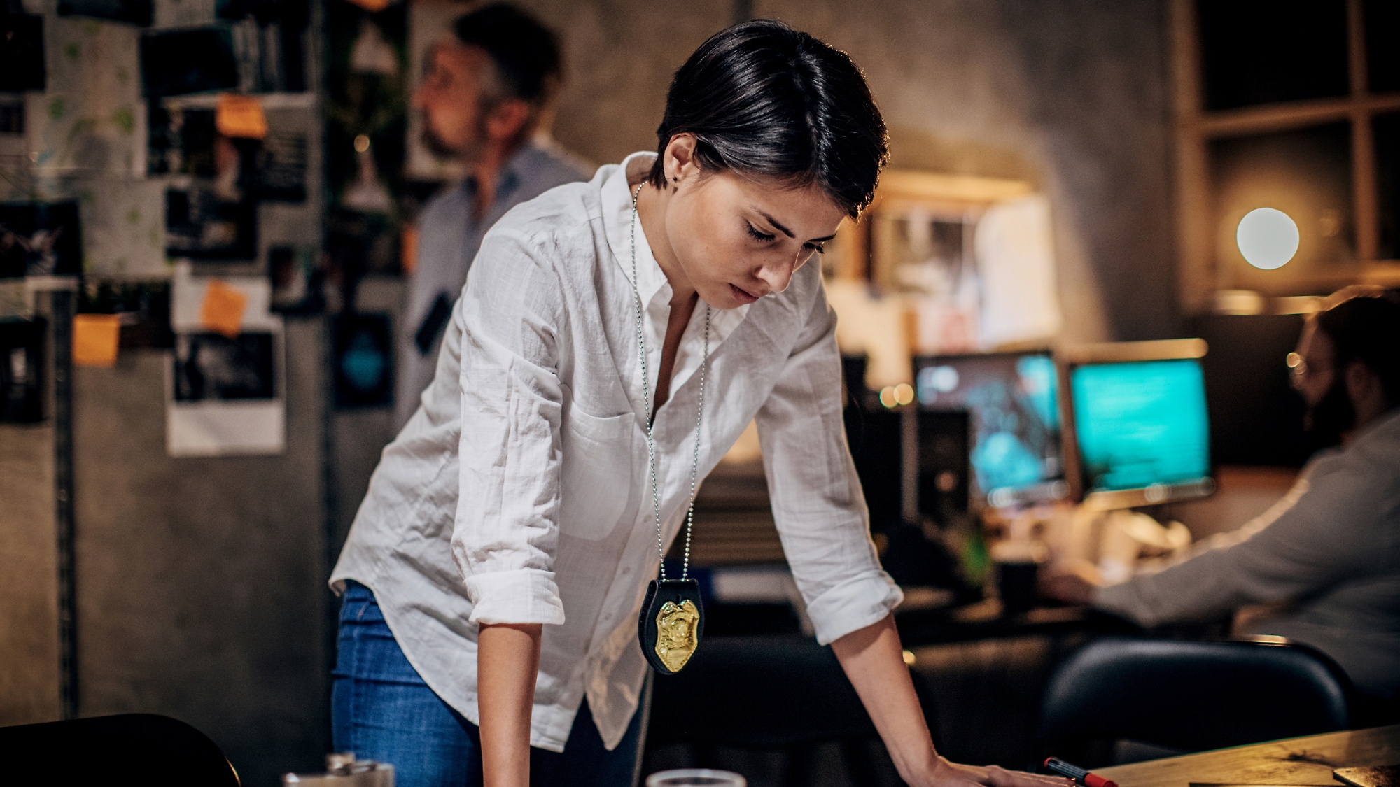 Una mujer detective concentrada con un distintivo se inclina sobre un escritorio en una oficina con poca iluminación y examina documentos