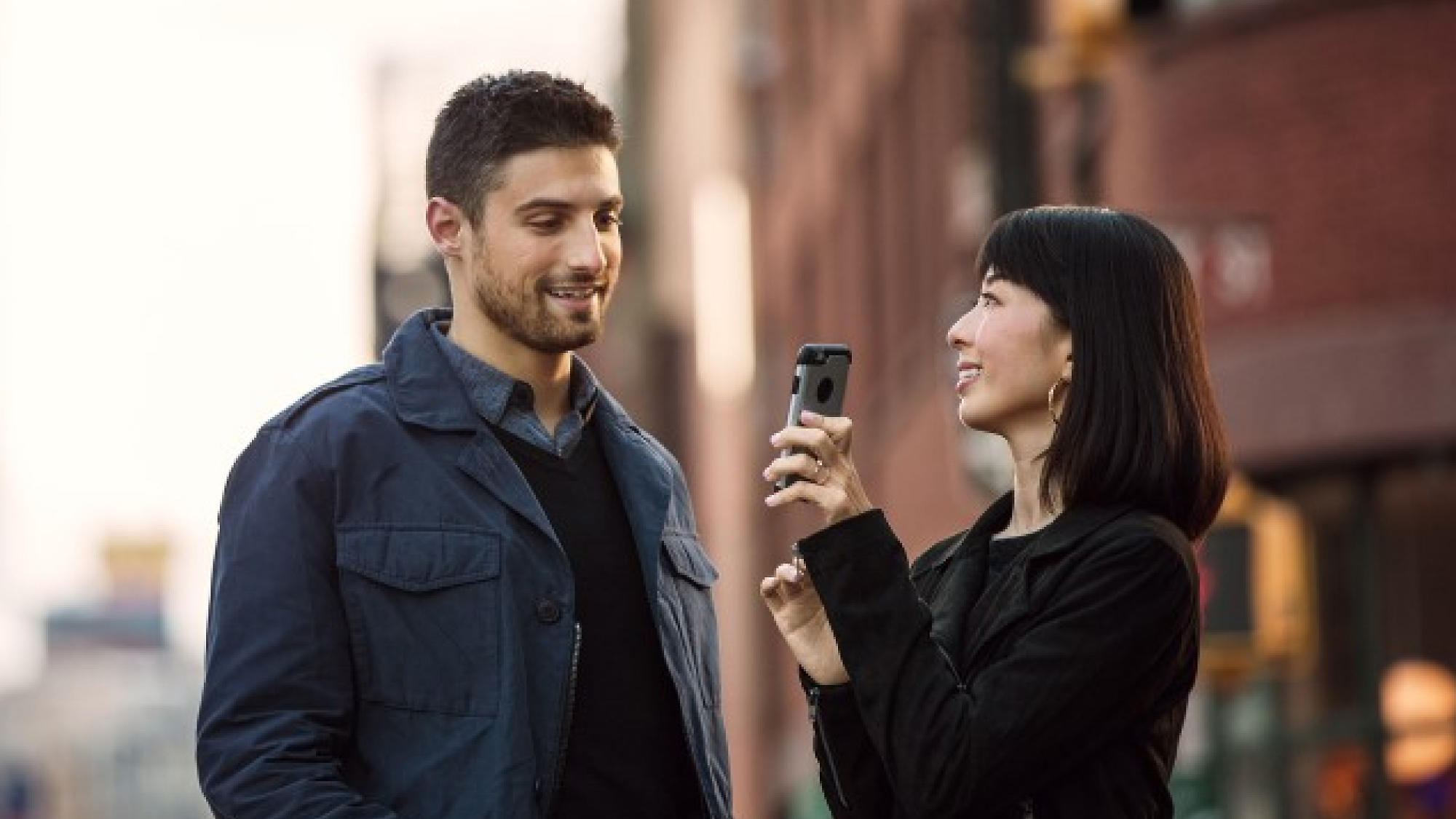 A woman holding a smartphone points it towards a man as they both stand on a city street, smiling and engaging in conversation.