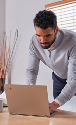 灰色のセーターを着た男性が、ブラインドのある明るく照らされたモダンな部屋で、木製の机の上のノート PC に身を乗り出して仕事をしています