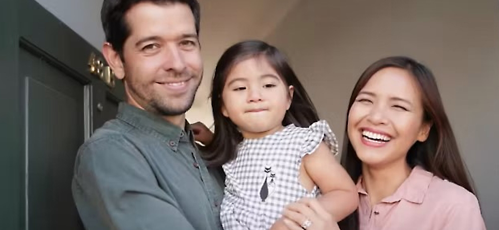 Famille heureux avec un homme, une femme et un jeune enfant souriant à l’appareil photo, debout à l’entrée d’une maison.