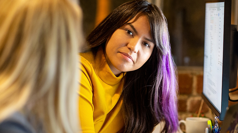 A woman in a yellow sweater attentively listens during a conversation in a casual office setting.