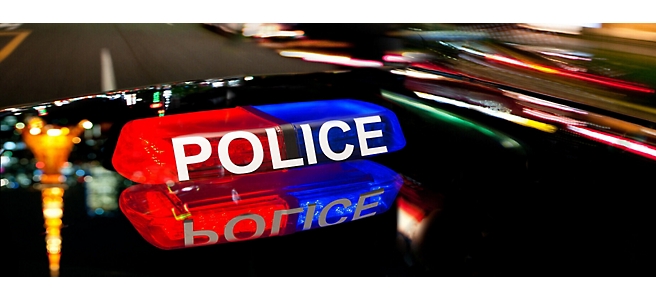 Voiture de police avec barre de toit lumineuse rouge, bleue et blanche sur fond estompé de rue de ville la nuit.
