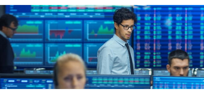 Uomo con occhiali e una maglietta in piedi in un ufficio di borsa occupato con dati finanziari su più schermi sullo sfondo.