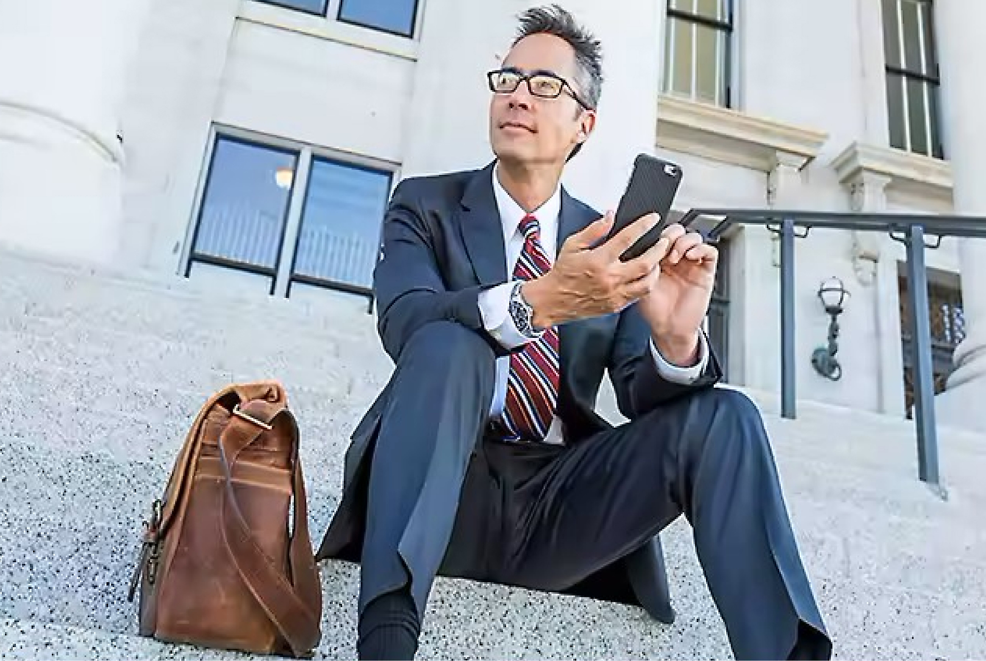 A businessman in a suit and glasses sits on steps outside a building, using a smartphone with a leather bag beside him.