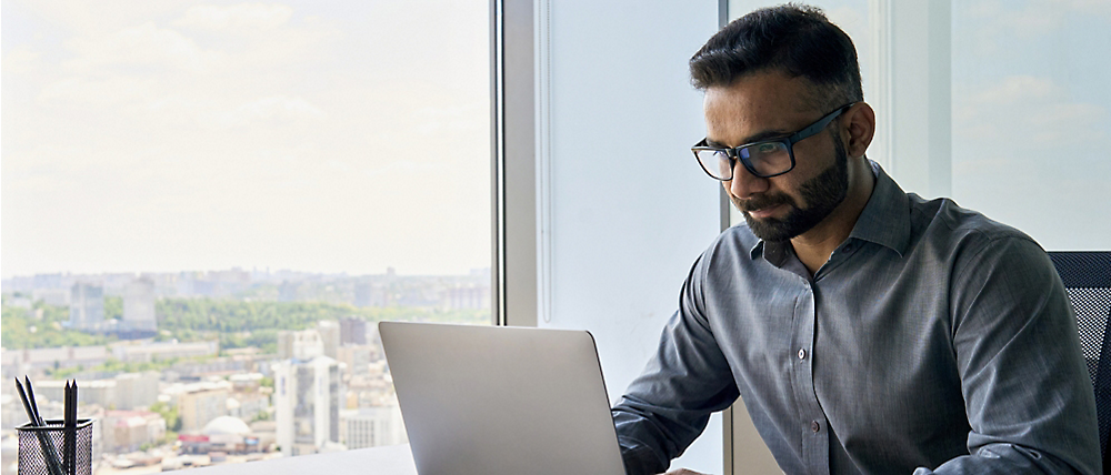 Pria berkacamata sedang bekerja pada laptop di kantor berskala tinggi dengan pemandangan kota di latar belakang.