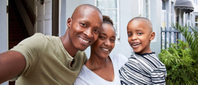 Nasmejana porodica – muškarac, žena i dečak – poziraju za selfi, stoje ispred svoje kuće.