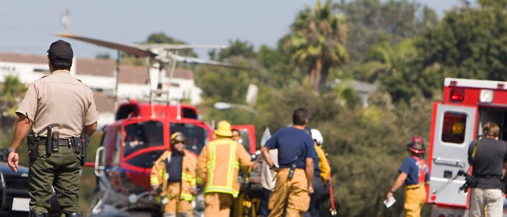 Emergency responders at a scene with a helicopter, ambulance, and firefighters, viewed from behind a sheriff's deputy.
