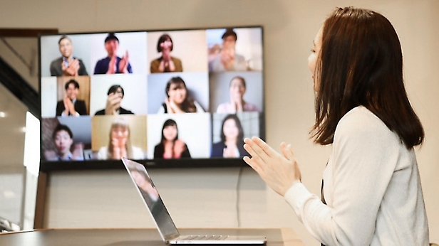 Mujer presentando a compañeros a través de una videoconferencia que se muestra en una pantalla grande en una oficina moderna.