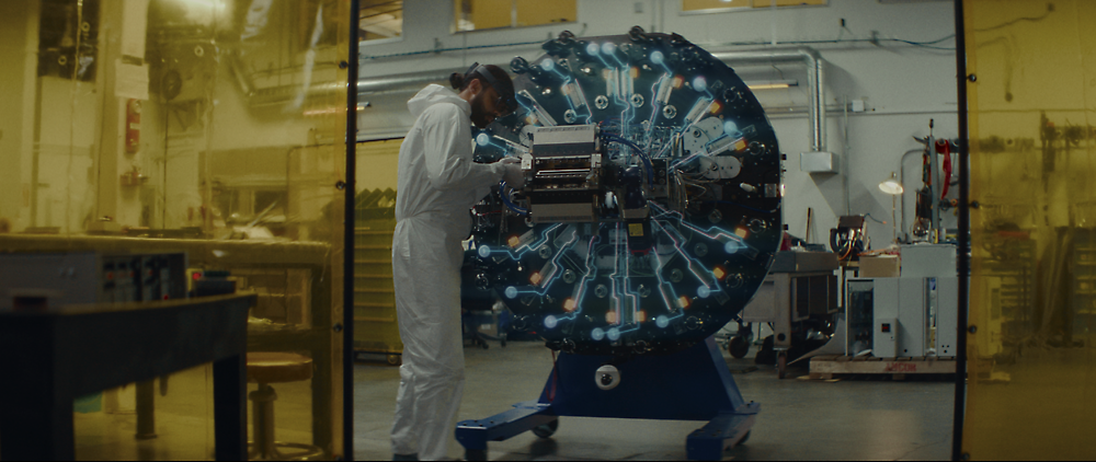 Uno scienziato con una tuta per camera sterile regola una macchina grande e complessa con cavi e luci blu luminose in un laboratorio high-tech.