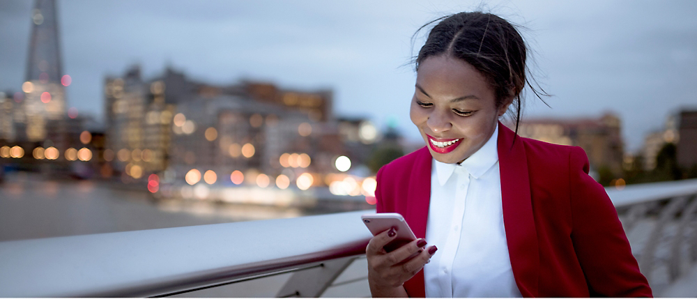 Una joven sonriente con una chaqueta roja usando su smartphone en un puente de la ciudad al atardecer.