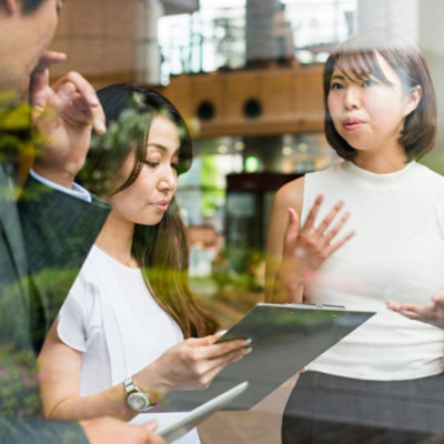 Two asian women and a man in a business meeting, one woman talking and gesturing, viewed through a glass window