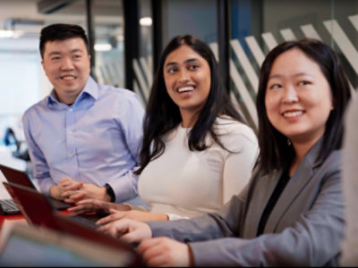 Four people in business attire are sitting at a conference table with laptops, engaging in conversation and smiling.