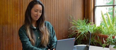 A woman with long hair sitting on a chair looking at a laptop on a desk in front of a window.