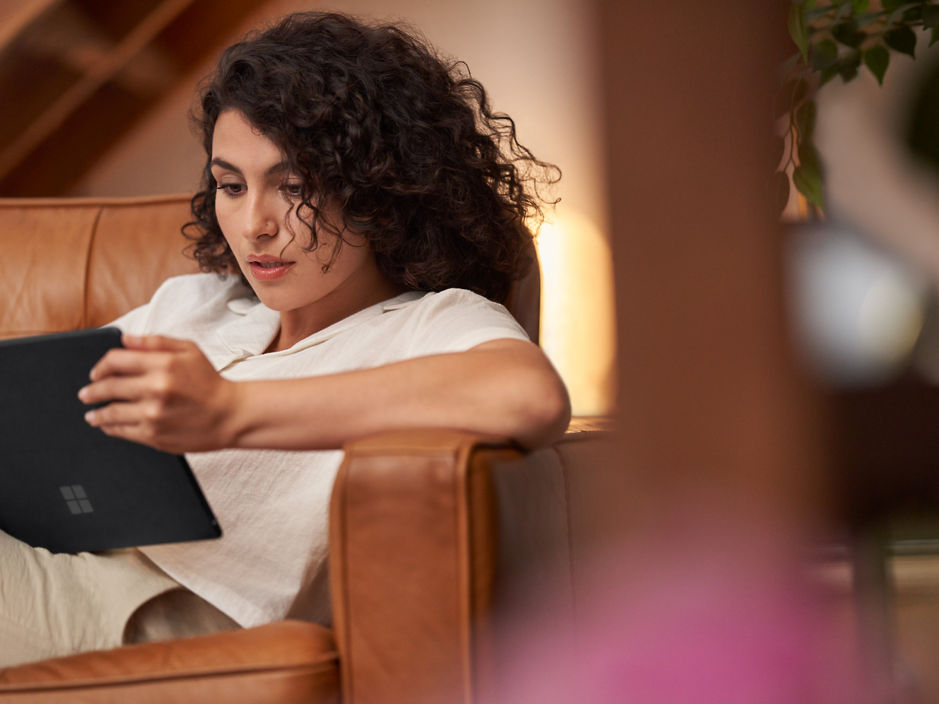 Mujer con el pelo rizado tratando de leer en una tableta mientras está sentada en un sillón de piel marrón interior.
