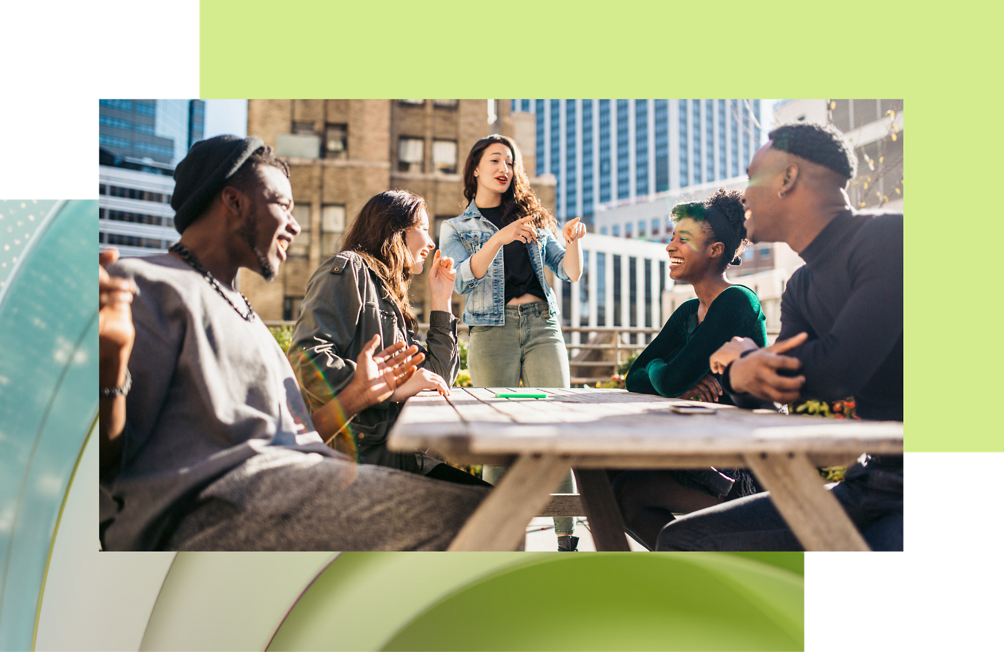 A diverse group of young adults enjoying a lively discussion around a table outdoors in a sunny urban setting.