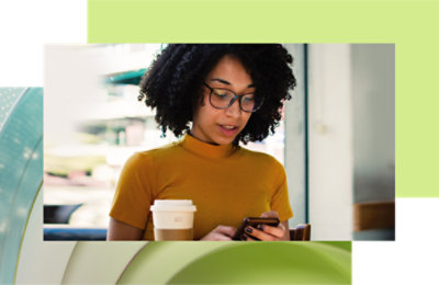 A woman in a yellow top and glasses using her smartphone while holding a coffee cup in a cafe setting.