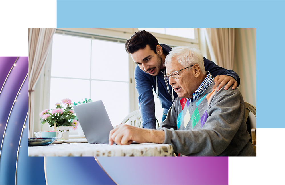 Young man assists elderly man with laptop in a bright room with flowers and curtains.