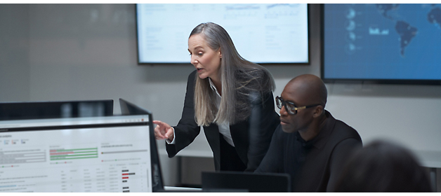Una mujer realiza una presentación con un hombre en una reunión, con pantallas digitales que muestran datos en segundo plano.