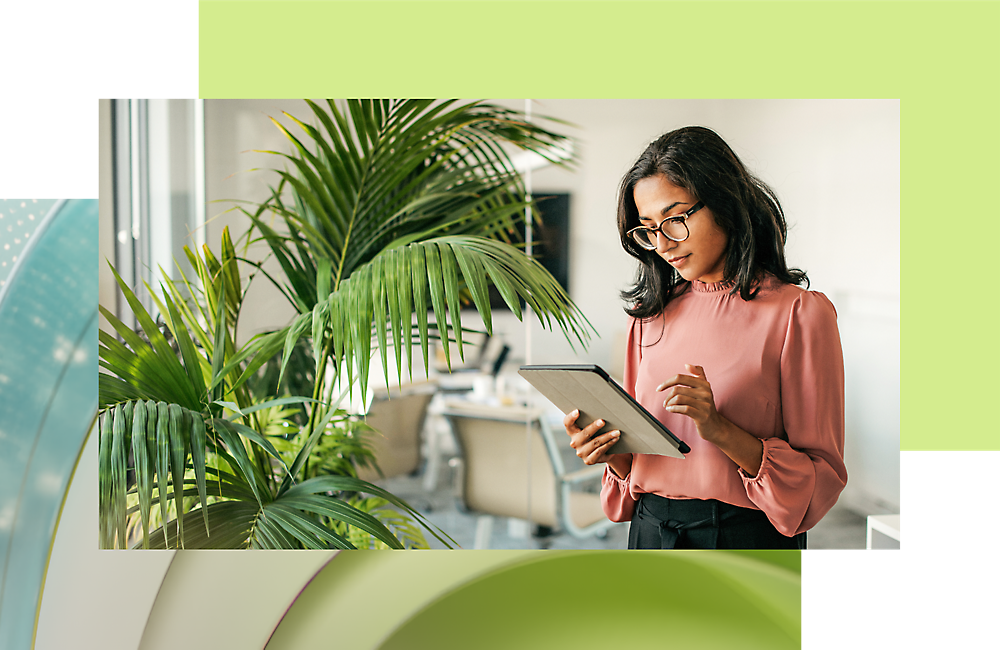 Gözlük takan ve pembe bluz giyen bir kadın, büyük pencereleri ve bitkiler olan modern bir ofiste bir tableti okuyor.