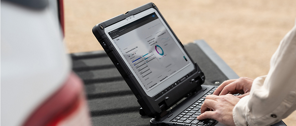 Una persona trabaja en un portátil resistente que muestra gráficos, sentado al aire libre en una superficie con arena.