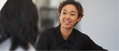 Person with short hair and a black shirt smile while talking to another individual, who is seen from behind.