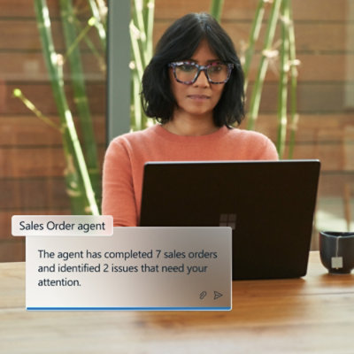 Egy szemüveges, rózsaszín pólós nő egy íróasztalnál ül egy laptop mellett.