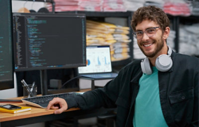 Smiling man with glasses and headphones sits at a desk with two monitors displaying code. Background shows shelves with packaged items.